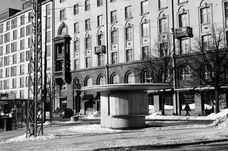A closed visor-roof kiosk in a snowy square beneath a tall apartment facade.
