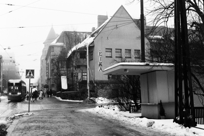 A tram approaches along a snowy street as a small kiosk sits to the right under trees.