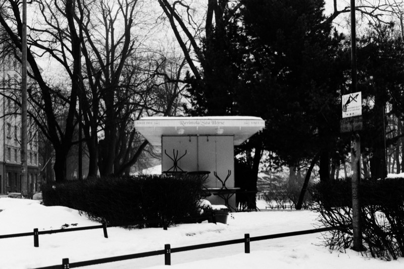 A shuttered kiosk in a snowy park, with stacked outdoor chairs under the roof.