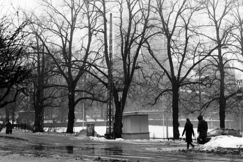A small kiosk beside bare trees on a wet winter street, with two people walking past.