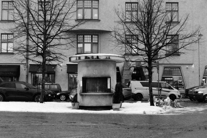 A compact round kiosk between two trees, with parked cars and shopfronts behind.