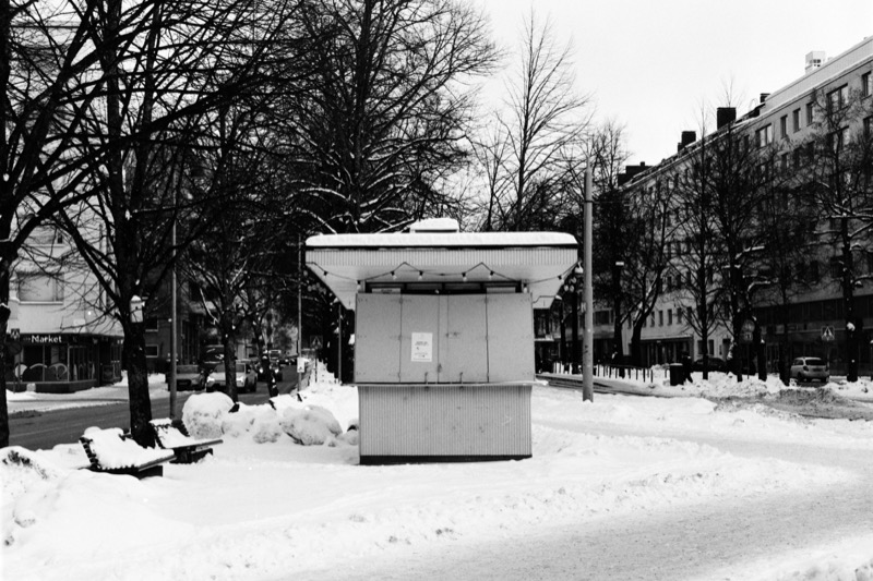 A closed kiosk centered in a snowy boulevard, with benches and apartment buildings on both sides.